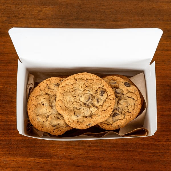 Three large chocolate chip cookies in a white box with brown paper, on a wooden table.